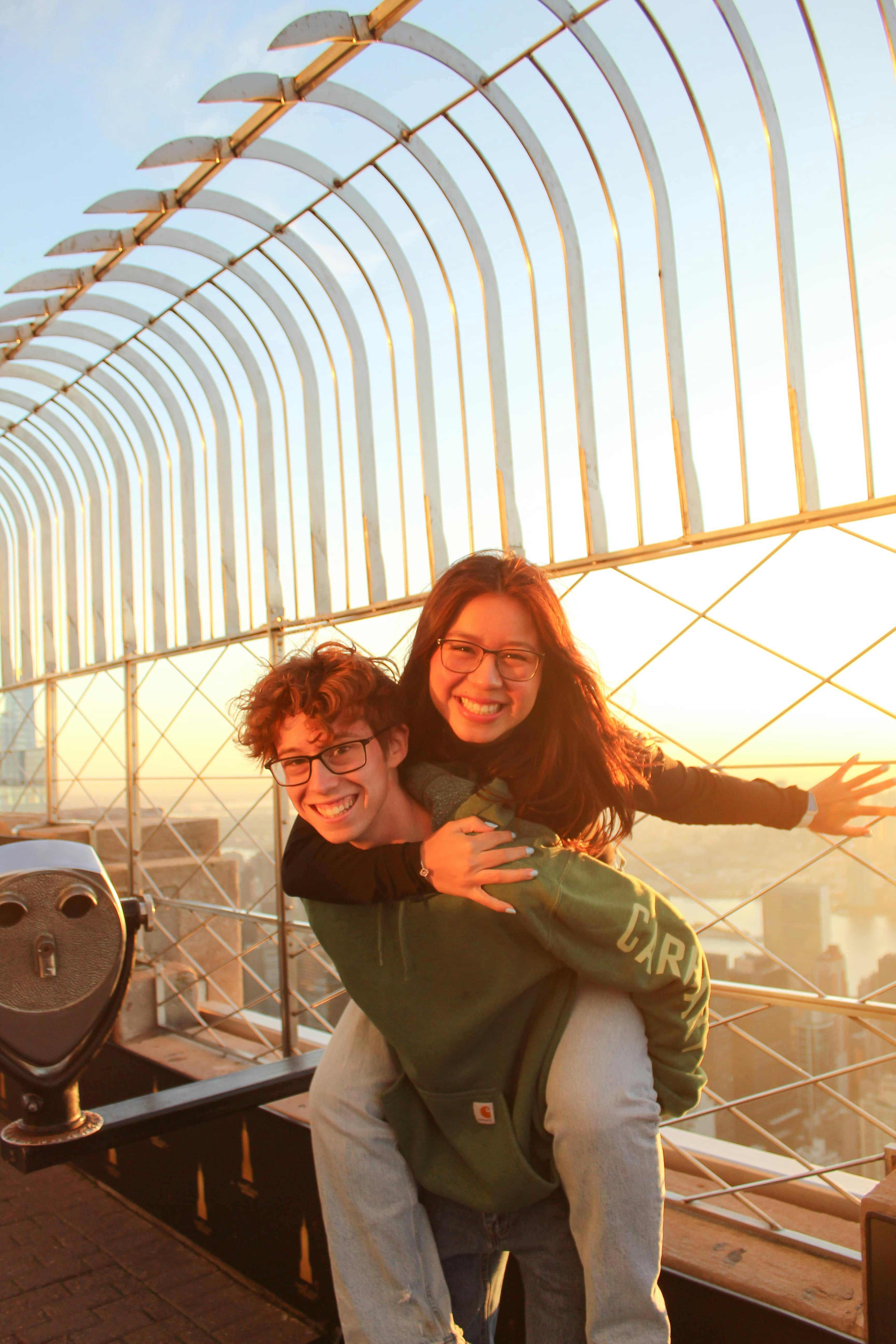 Two people smiling at sunset on a viewing platform, with one giving a piggyback ride and cityscape visible in the background.