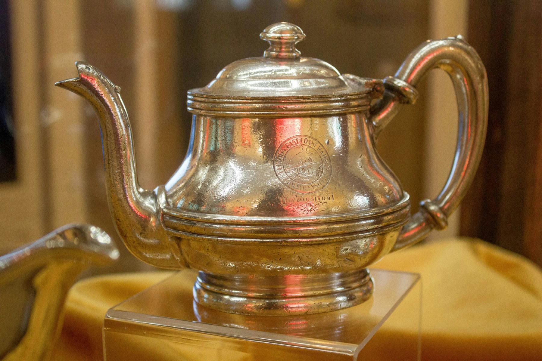 En polerad tekanna i silver med invecklade handtag och ett lock som visas på ett genomskinligt stativ. Gravering synlig på sidan.
