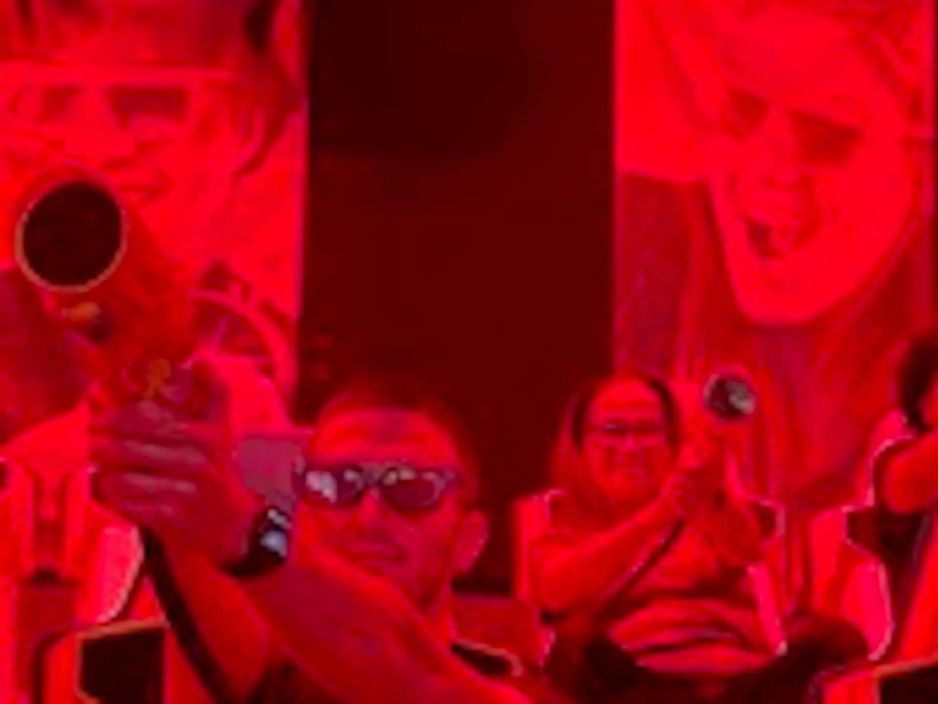 People wearing 3D glasses sit in red seats, holding game controllers, immersed in a dark, red-lit gaming environment.