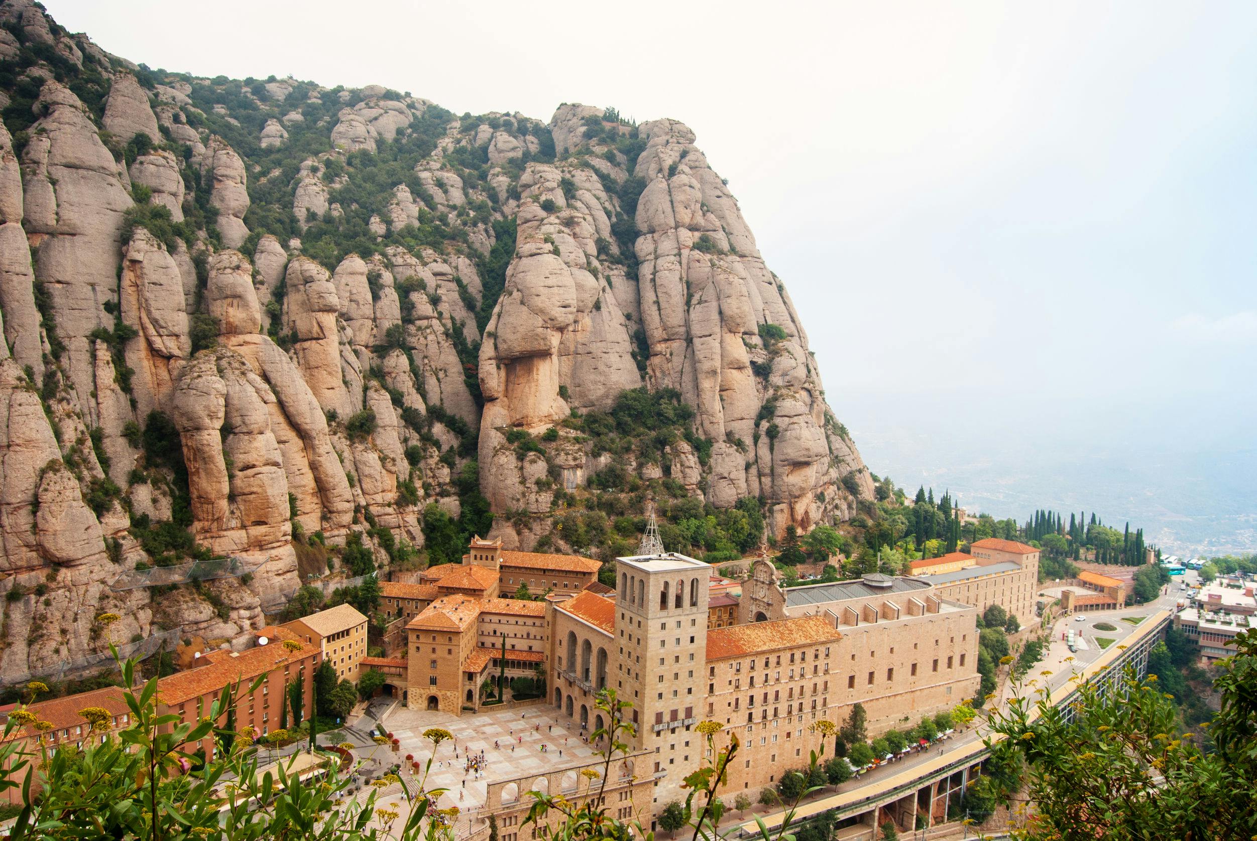 Historic monastery with terracotta roofs situated at the base of a massive, rocky mountain range under a hazy sky.