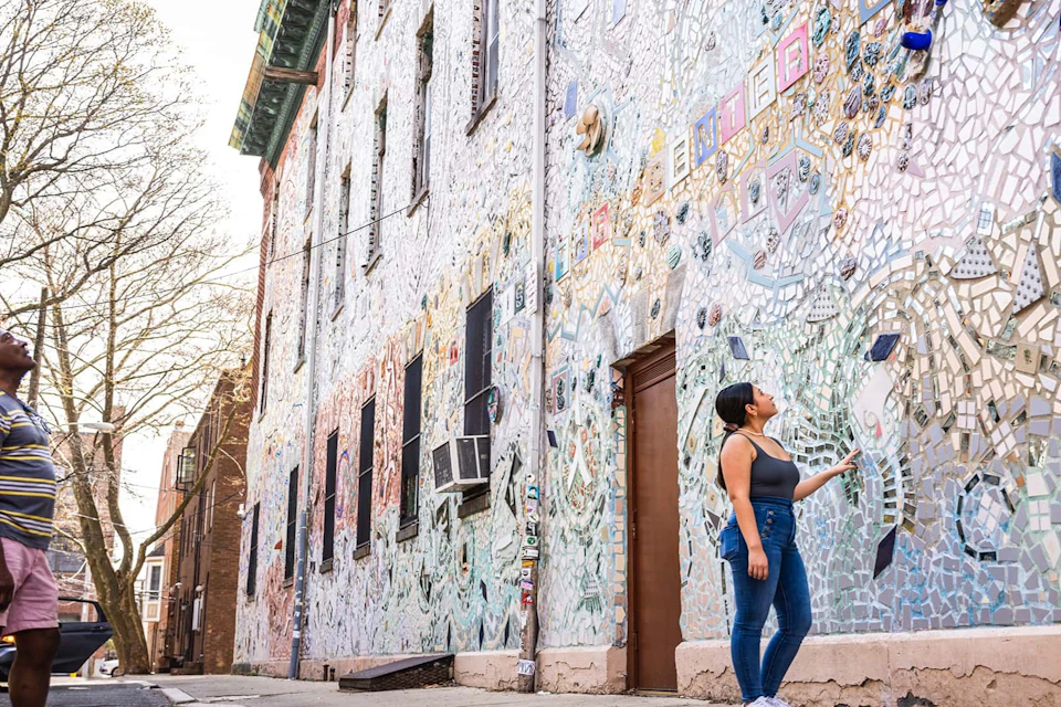A woman stands and admires a building covered in colorful mosaic artwork on a sunny day.