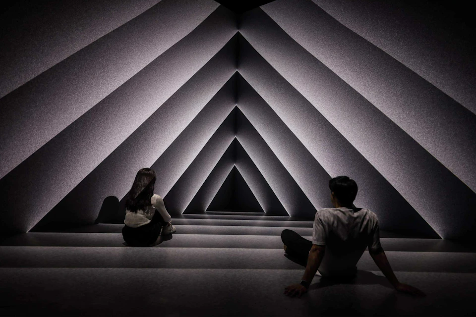 Two people sit on a geometric, triangularly patterned structure illuminated by soft lighting, creating a tunnel effect.
