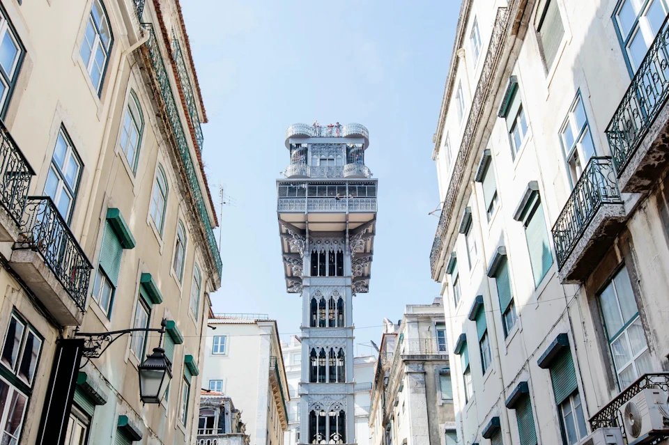 A historic iron elevator tower stands between beige and white buildings under a clear, blue sky.