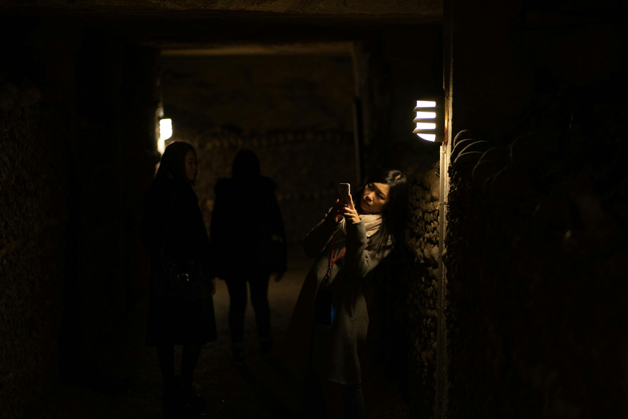 Three people in a dimly lit, narrow corridor, with one woman taking a photo with her phone beneath a wall light.