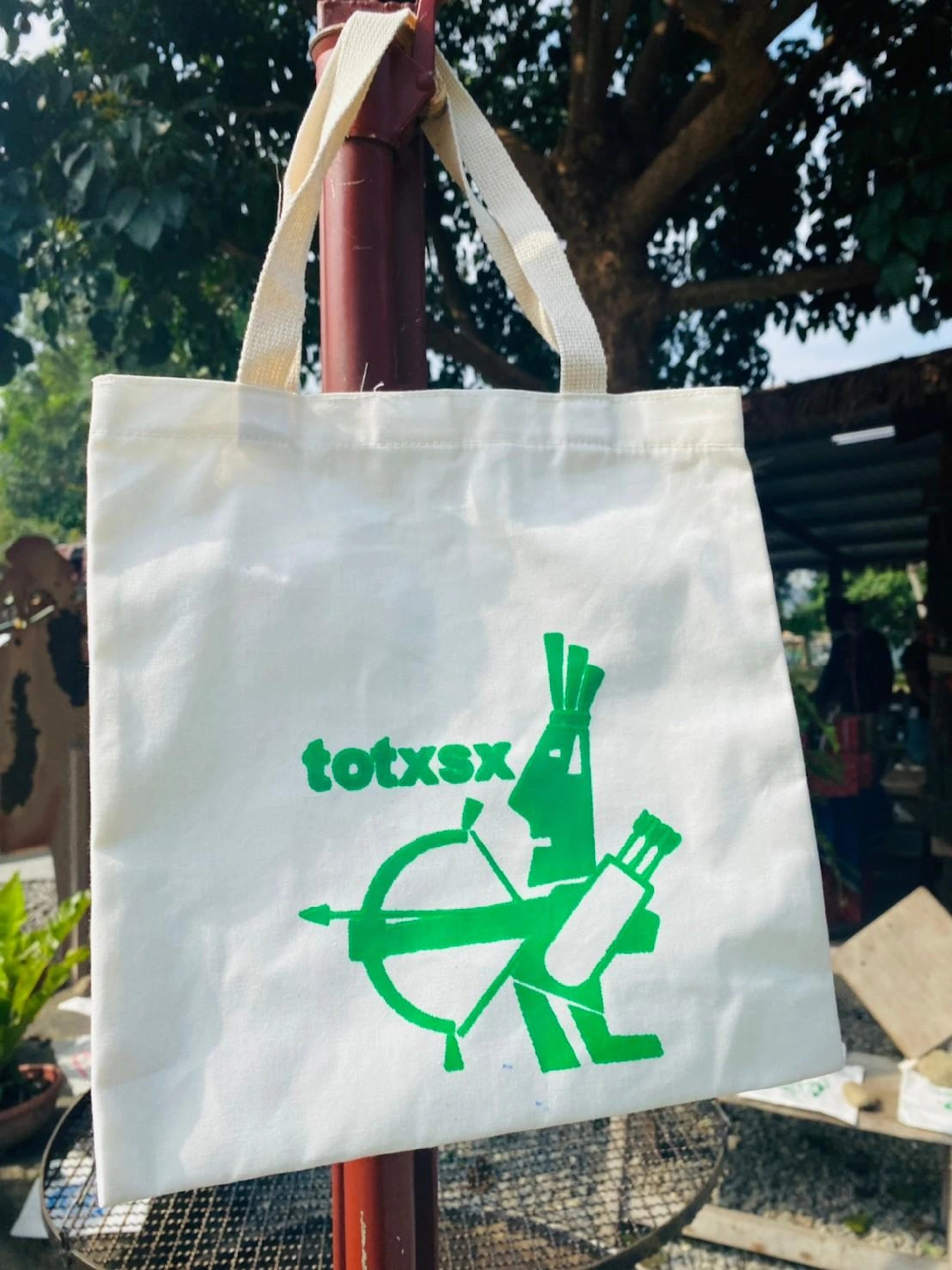 A white tote bag with a green graphic design and text "totxsx," hanging outdoors near a tree.