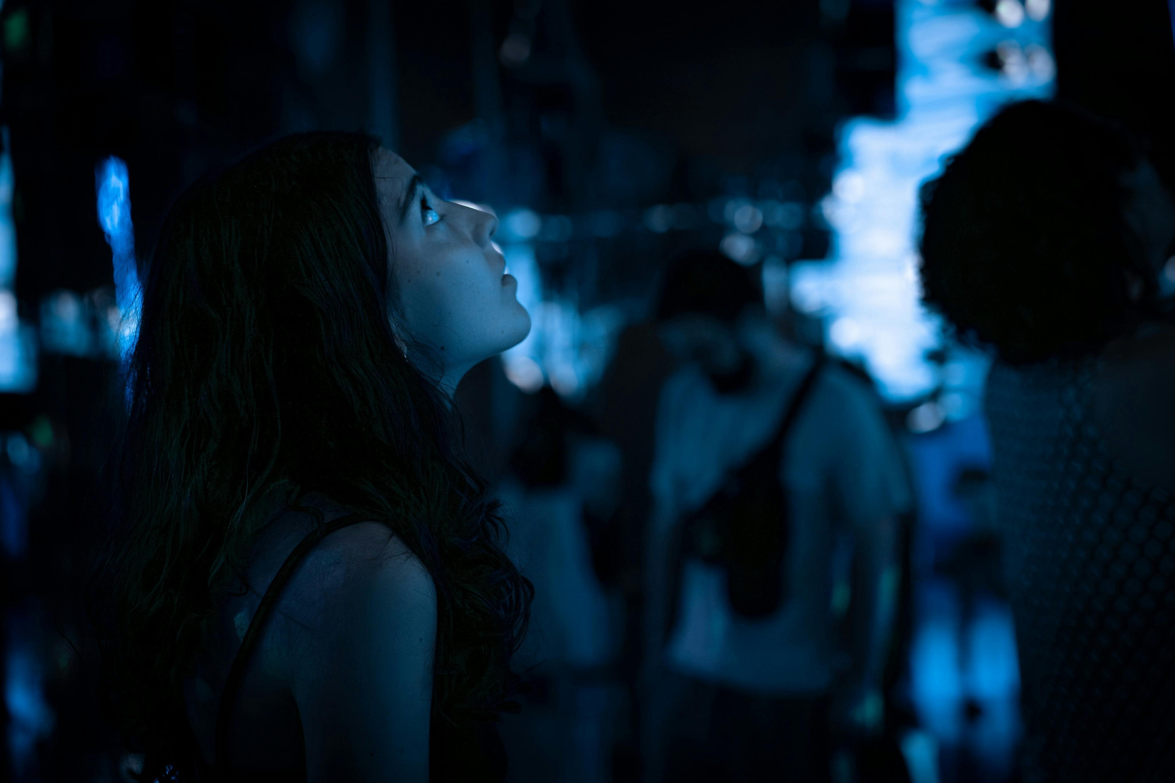 Žena vzhlédne v tlumeně osvětlené místnosti s modrým osvětlením; lidé jsou rozmazaní v pozadí.
