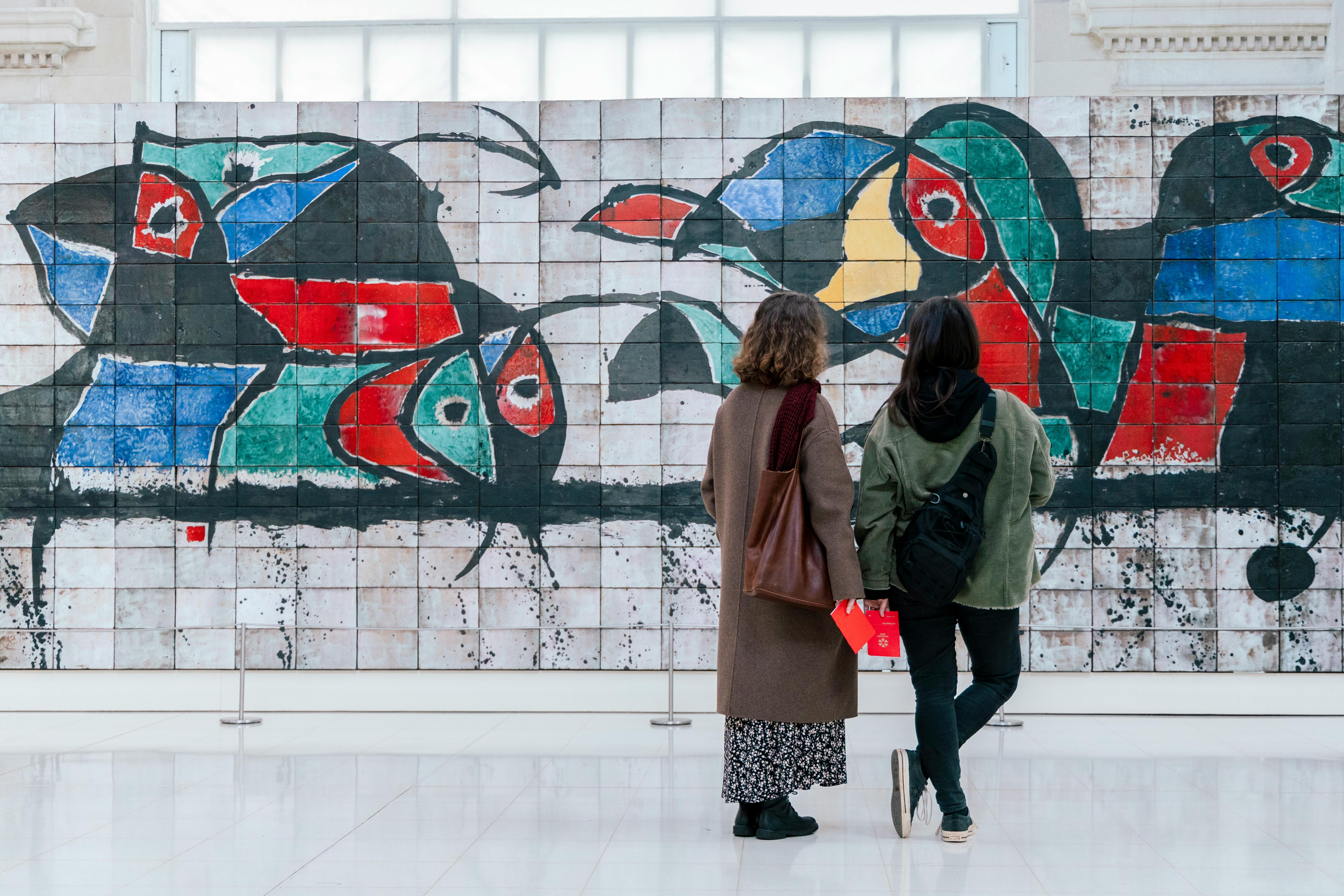 Deux personnes se tiennent devant une peinture murale abstraite et colorée, composée de carreaux de faïence, dans un espace intérieur lumineux.