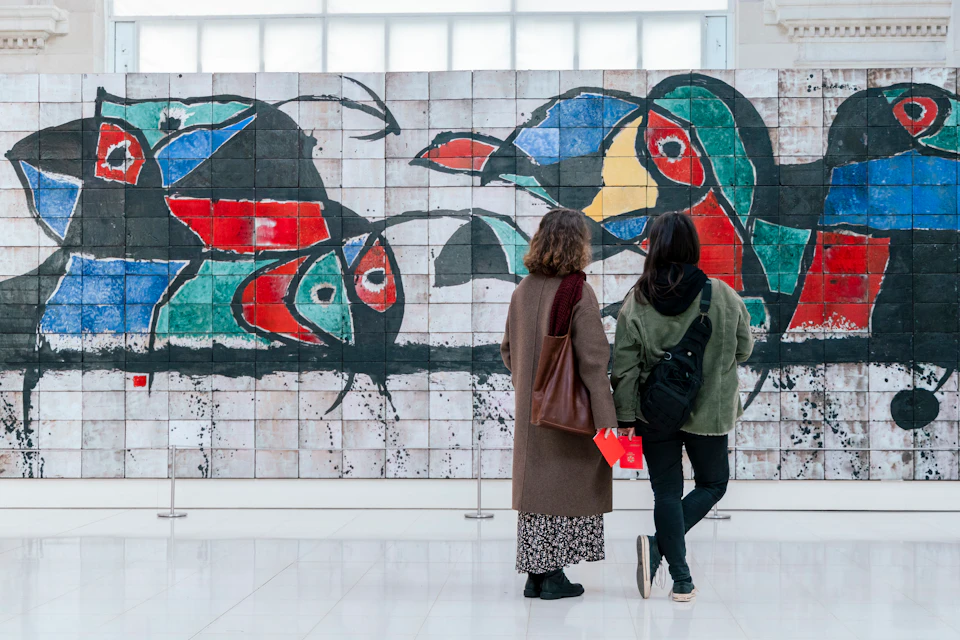 Two people stand in front of a colorful abstract mural composed of tiled artwork in a bright indoor space.