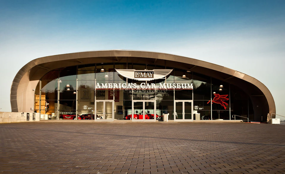 Exterior of LeMay - America’s Car Museum with a curved glass facade and visible classic cars inside.
