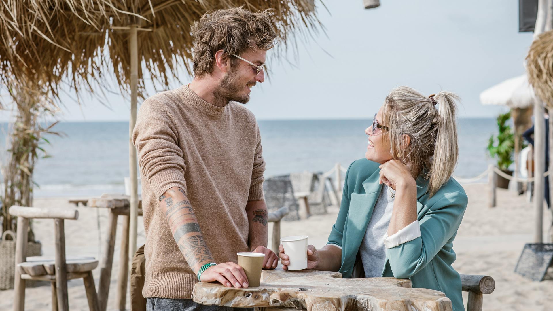 To mennesker, der smiler og holder drinks, står ved et træbord på en strand med havet i baggrunden.