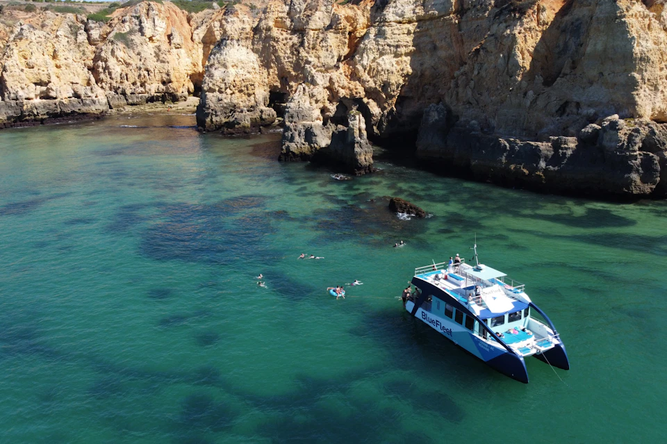 A catamaran with "BlueFleet" branding floats in clear, green water near rocky cliffs; people swim nearby.
