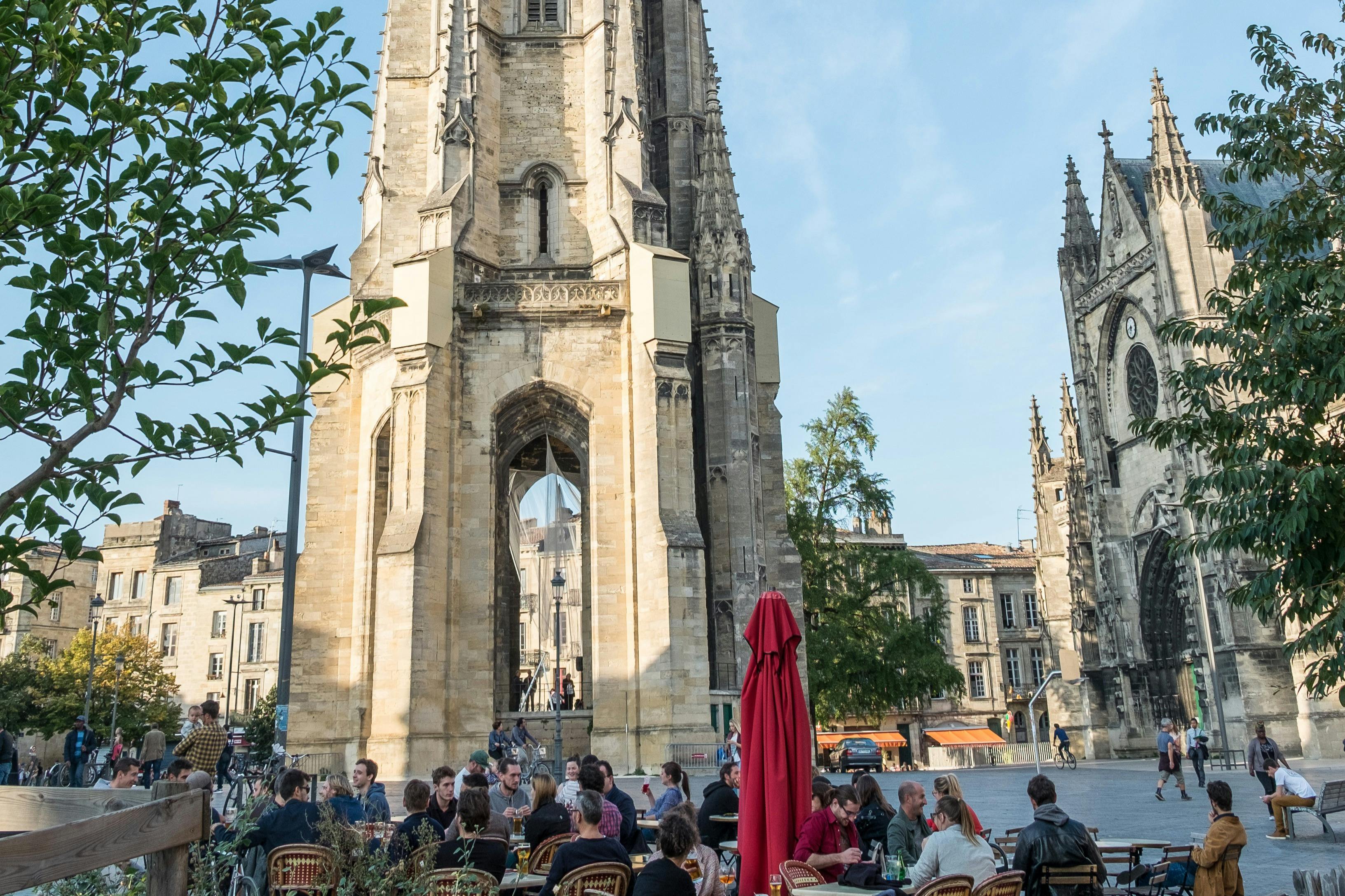 Människor sitter på ett uteservering nära ett högt stentorn och historiska byggnader på ett soligt stadstorg.