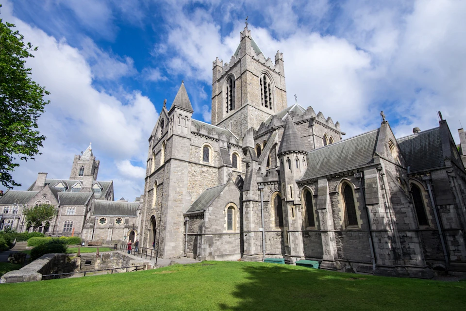 Stone cathedral with pointed arches, tall tower, and multiple spires under a partly cloudy sky, situated next to a lawn.