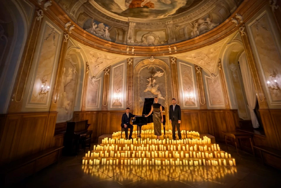 Three people stand near a piano surrounded by numerous lit candles in a grand hall with ornate murals and arches.