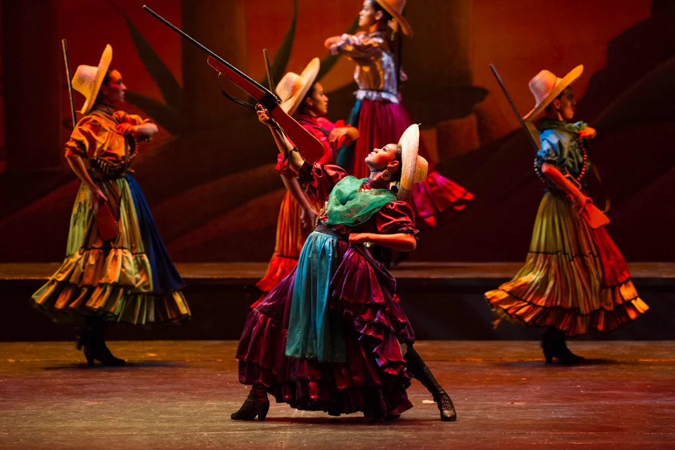 Dancers in colorful traditional dresses and wide-brimmed hats perform on stage, one holding a rifle aloft.