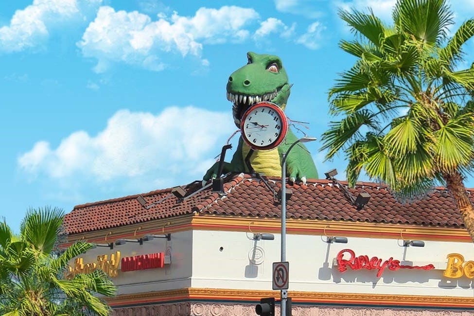 一尊巨大的绿色恐龙雕像打破了建筑物的屋顶，前面的一根柱子上挂着一个时钟，周围环绕着棕榈树。