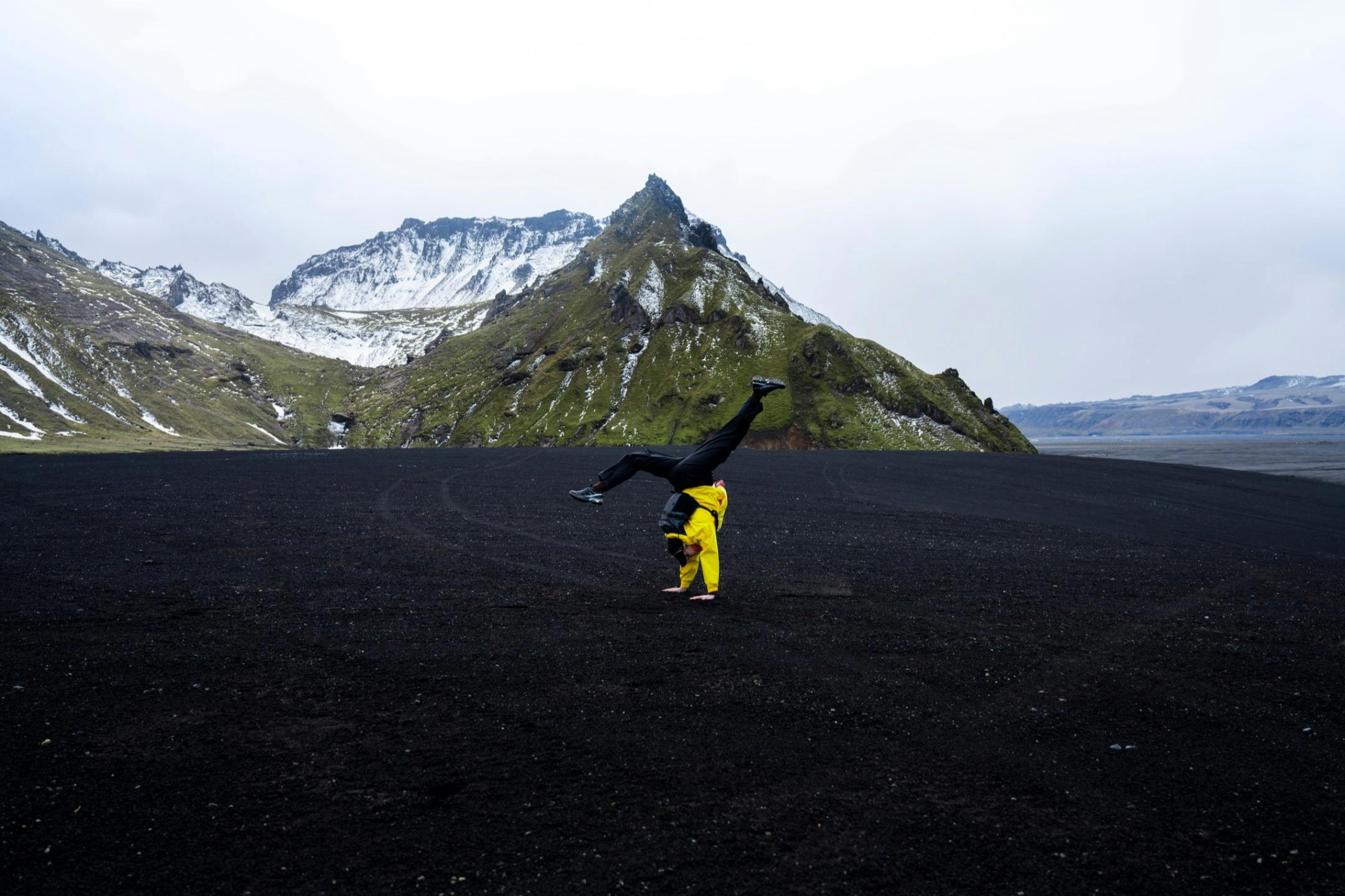 A person in a yellow jacket performs a handstand on a dark plain with a green and snowy mountain in the background.