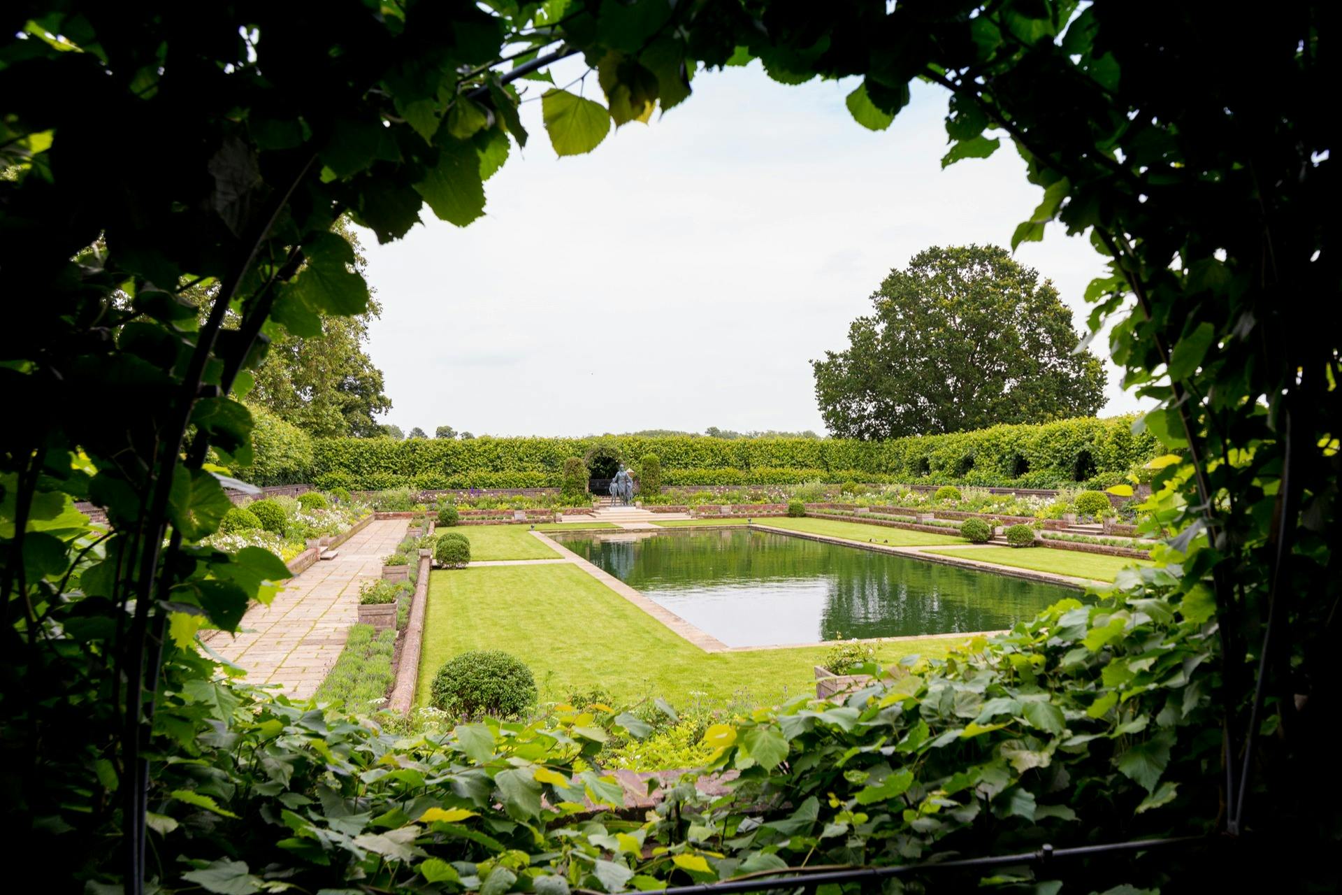 A symmetrical garden with a central rectangular pond, surrounded by hedges and greenery, viewed through a leafy arch.