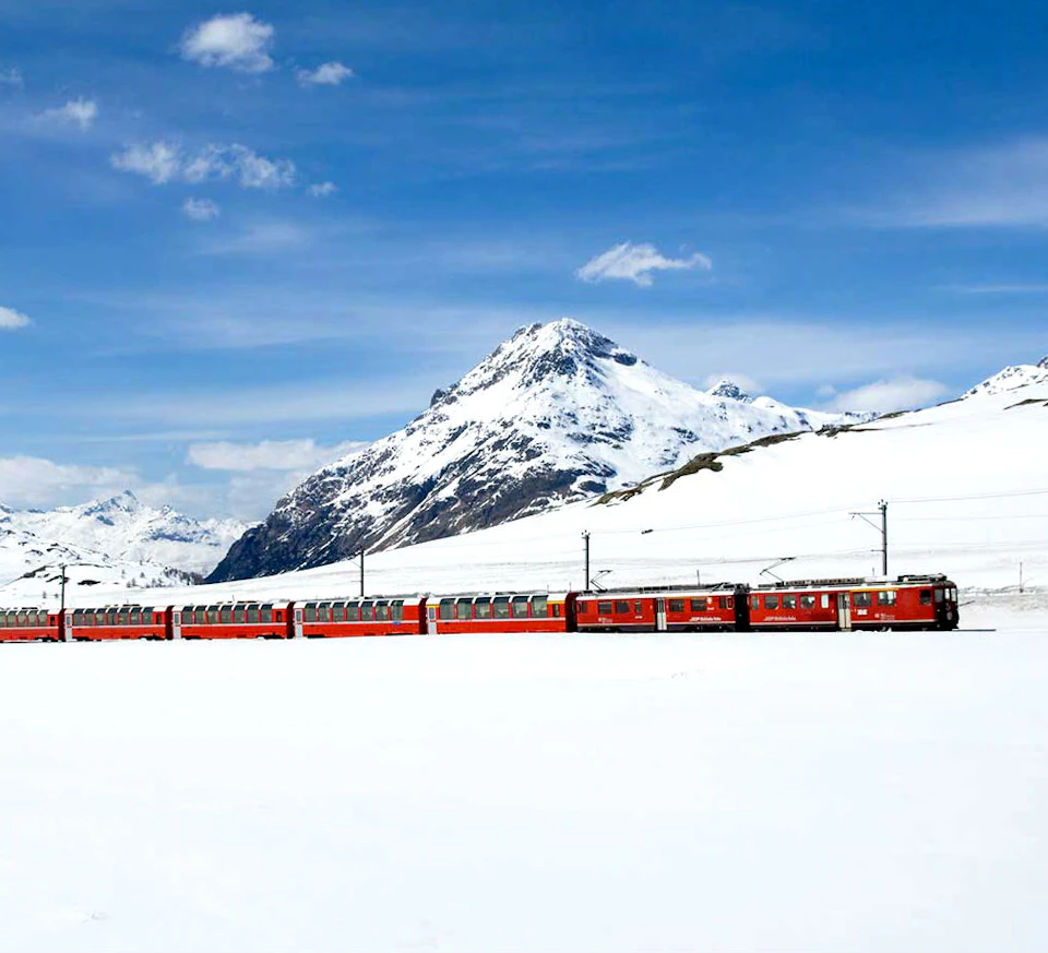 A red train travels through a snowy landscape with a towering, snow-covered mountain and a blue sky in the background.