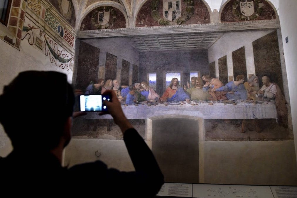 A person photographs the painting 'The Last Supper' by Leonardo da Vinci, displayed on a wall in a museum setting.