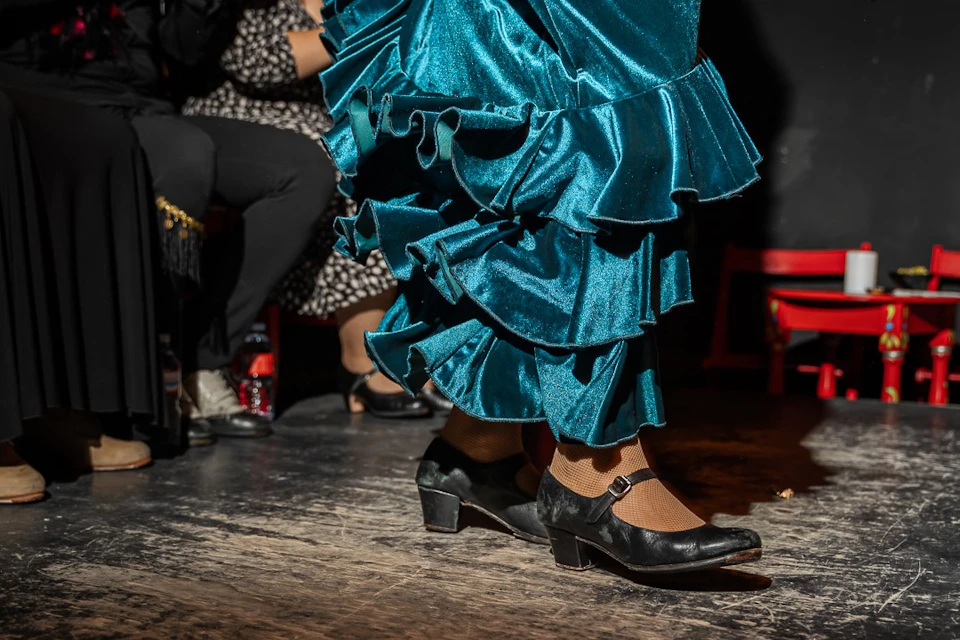 A person wearing a teal ruffled dress and black heeled shoes is dancing on a wooden floor, with seated spectators in the background.