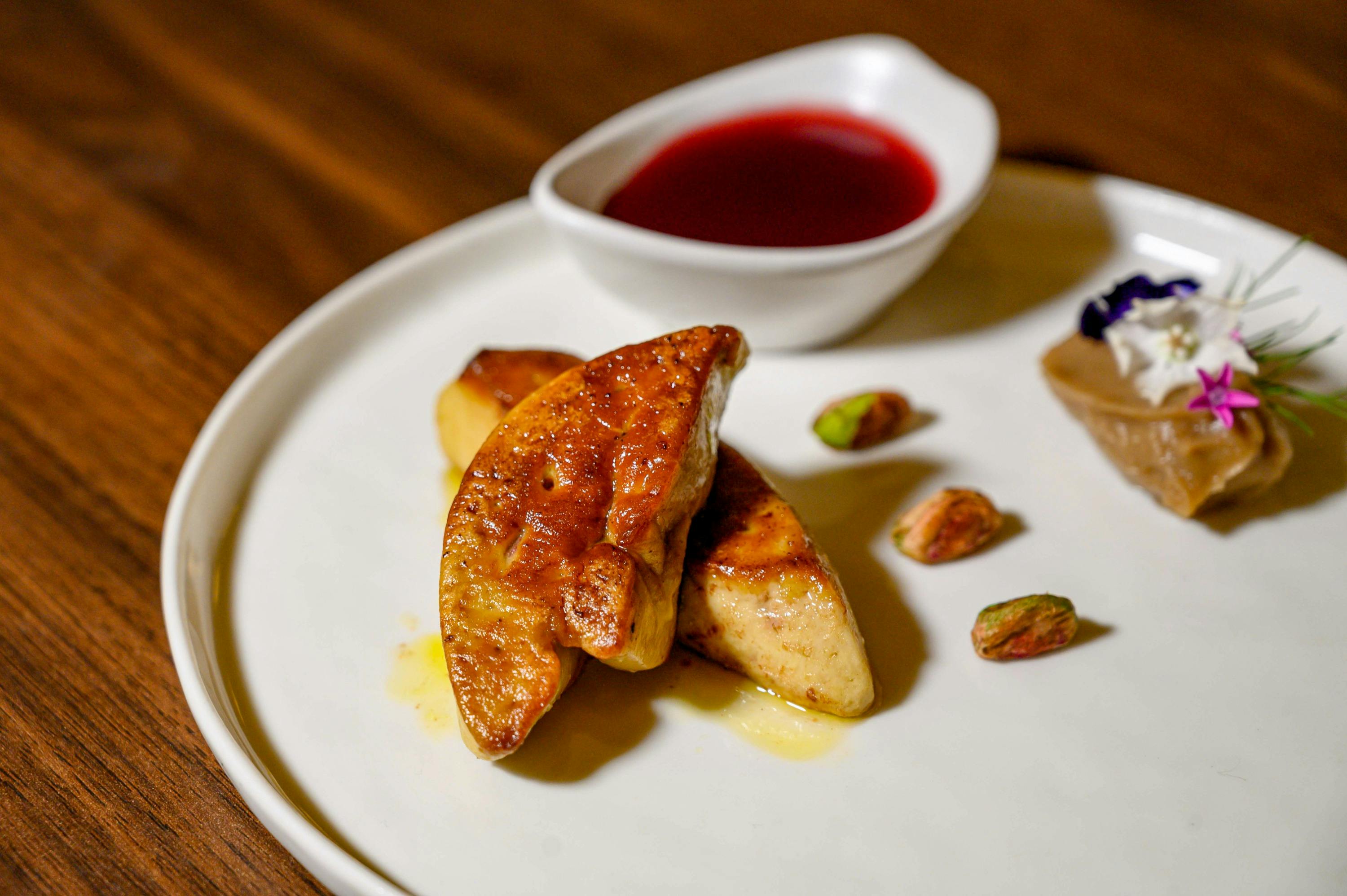 Seared foie gras served on a white plate with a small bowl of red sauce and pistachios as garnish.