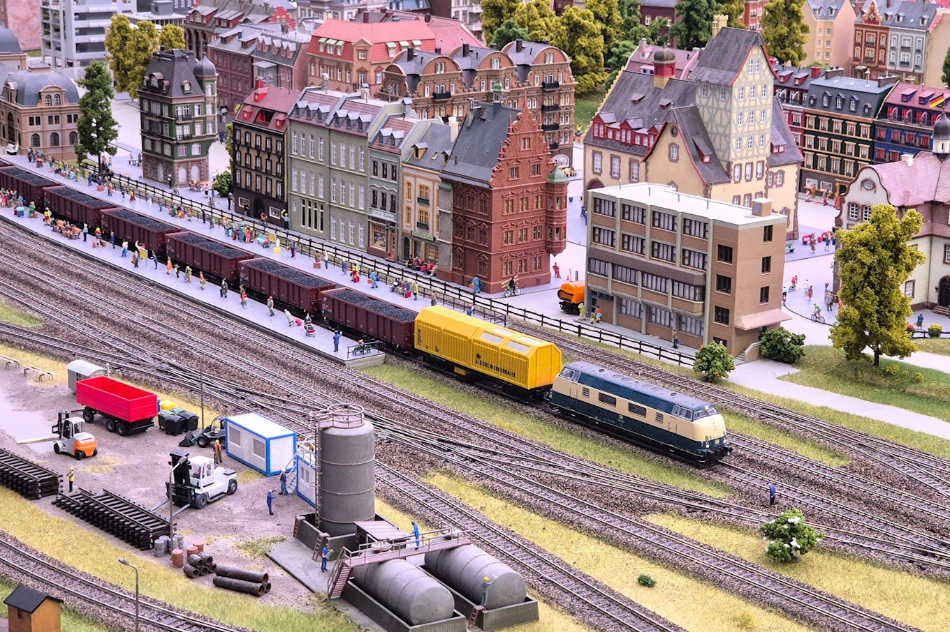 A miniature model village featuring a train station, detailed buildings, trains on tracks, and small human figures on the platform.