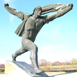 En stor utomhusbronsstaty av en person i en dynamisk löpställning, som håller ett långt föremål ovanför, mot en lantlig bakgrund.