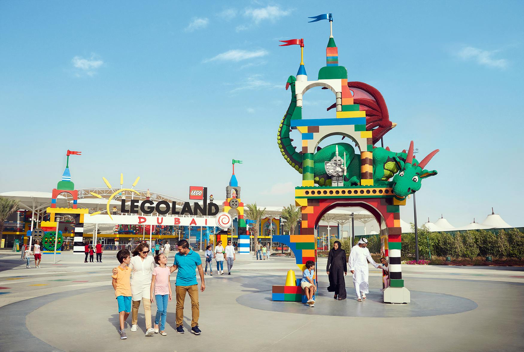 Familien spazieren in der Nähe des farbenfrohen Eingangs von LEGOLAND Dubai, der einen großen LEGO Drachen und einen lebhaften Torbogen unter einem klaren Himmel zeigt.