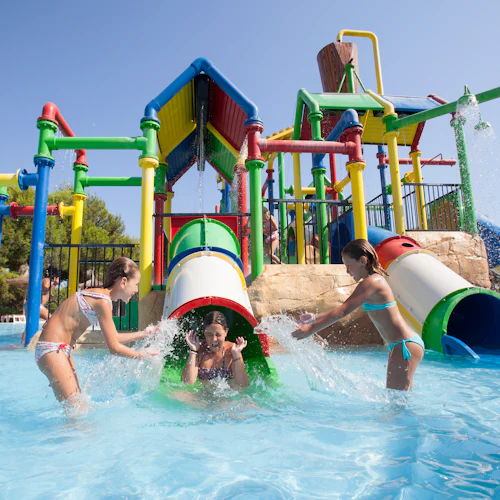 Drei Kinder spielen in einem Wasserpark mit bunten Rutschen und Wasserspritzern unter einem strahlend blauen Himmel.