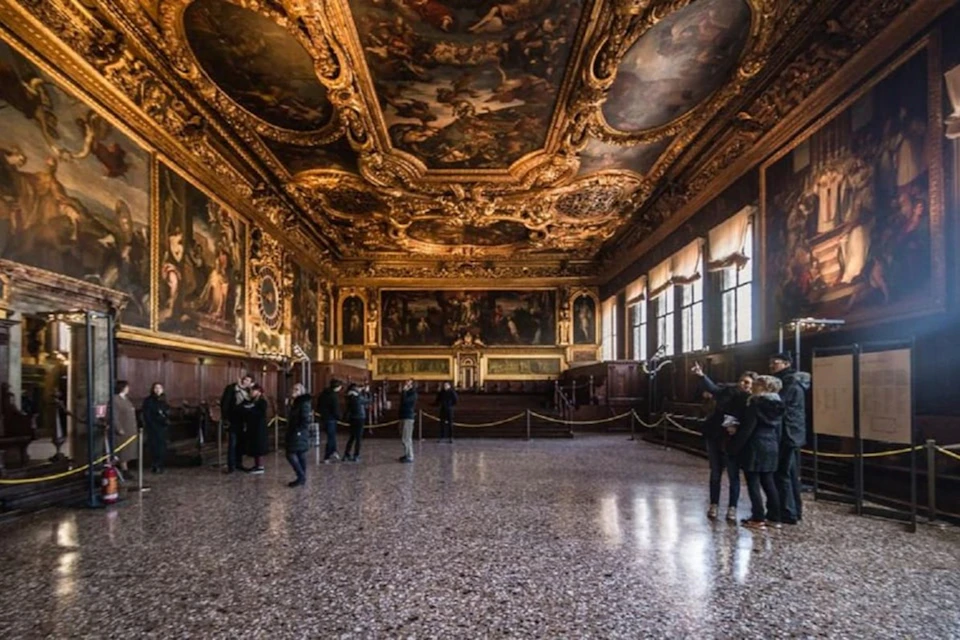 A grand, ornate hall with a gilded ceiling, large paintings on the walls, and people observing the space.