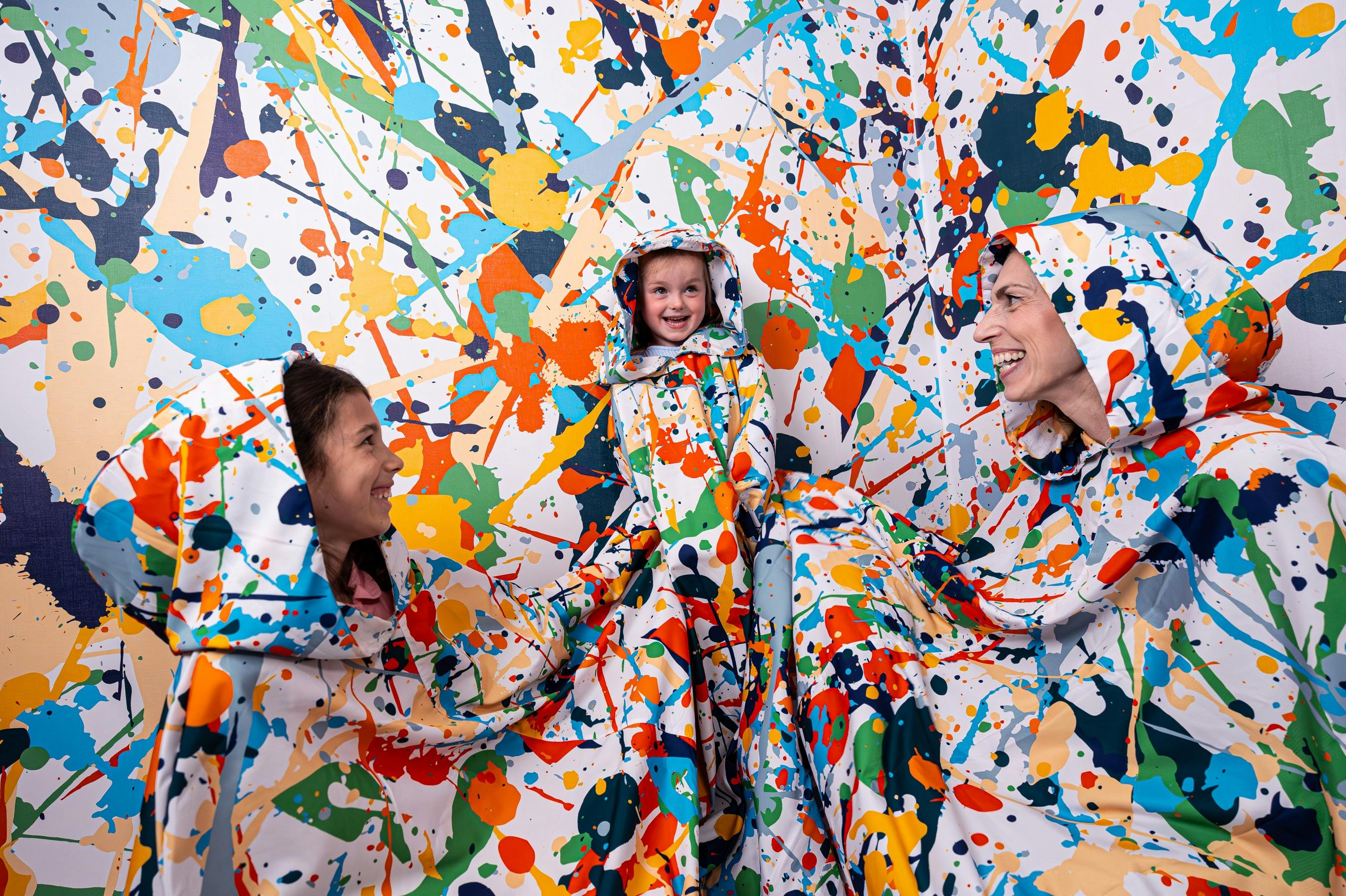 Tre personer bär färgglada stänkmönstrade kappor och ler i ett rum med matchande stänkmönstrade väggar.