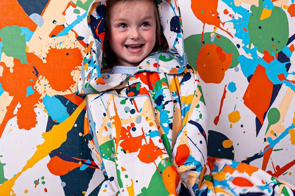 Three people wearing colorful splatter-patterned cloaks, smiling in a room with matching splatter-patterned walls.