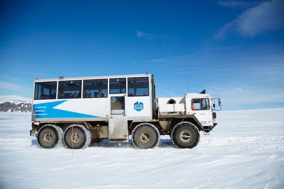 A large off-road vehicle with "Into the Glacier" branding drives on snow, carrying passengers under a clear blue sky.