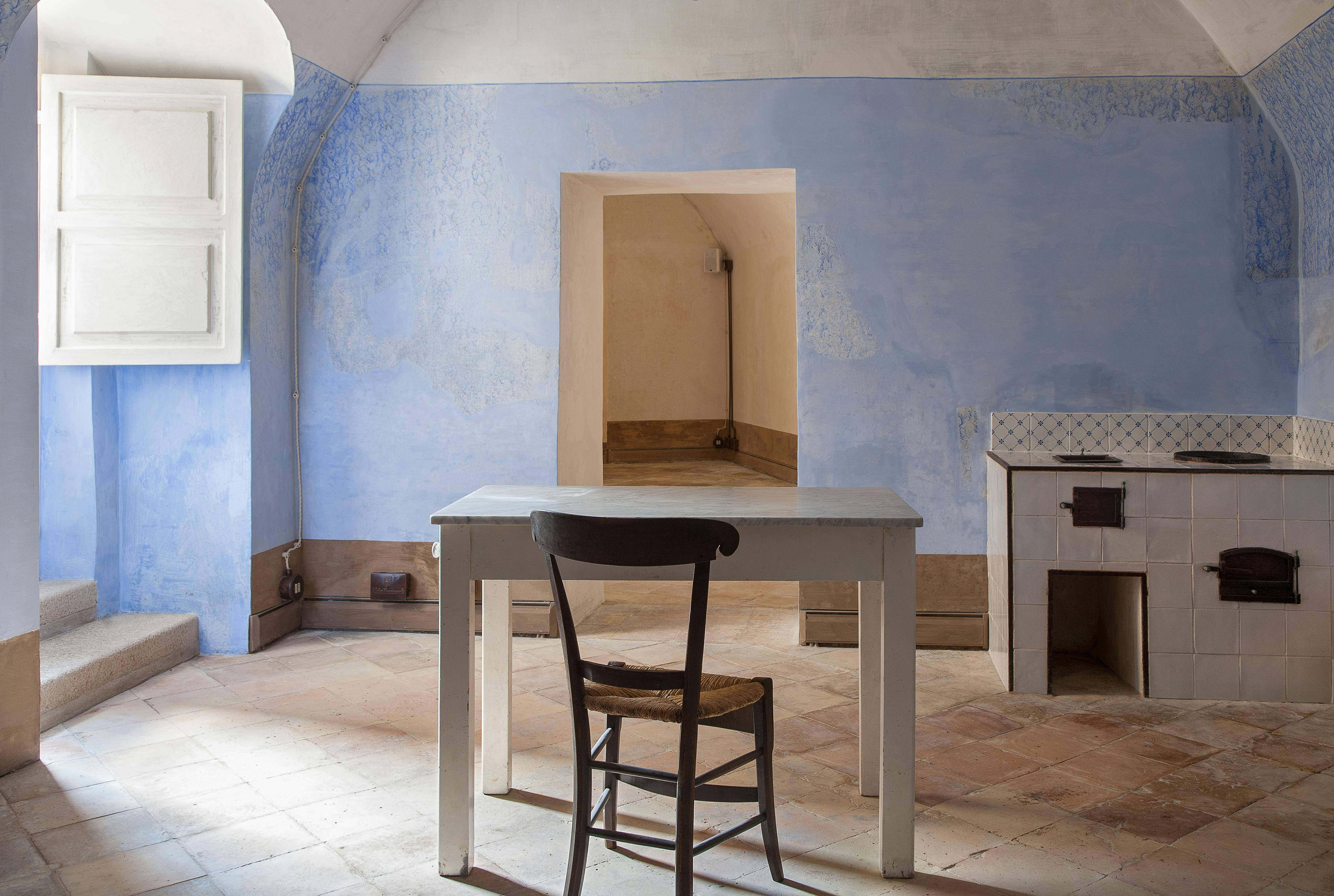 A simple room with light blue walls, a white table, and a wooden chair on tiled flooring. An open doorway and window are visible.