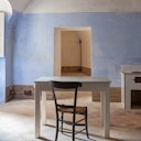 A simple room with light blue walls, a white table, and a wooden chair on tiled flooring. An open doorway and window are visible.
