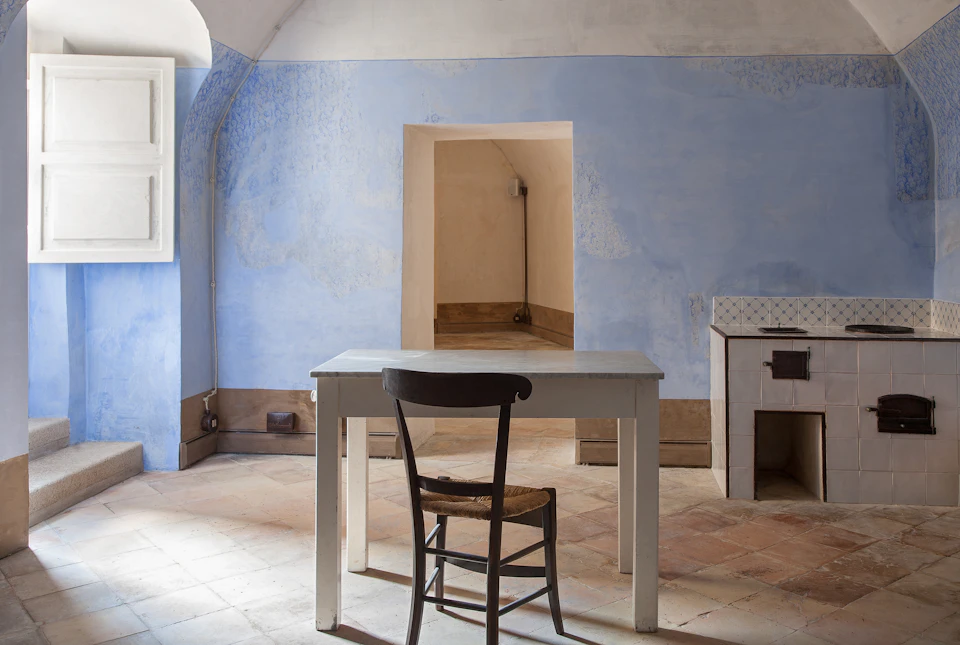 A simple room with light blue walls, a white table, and a wooden chair on tiled flooring. An open doorway and window are visible.