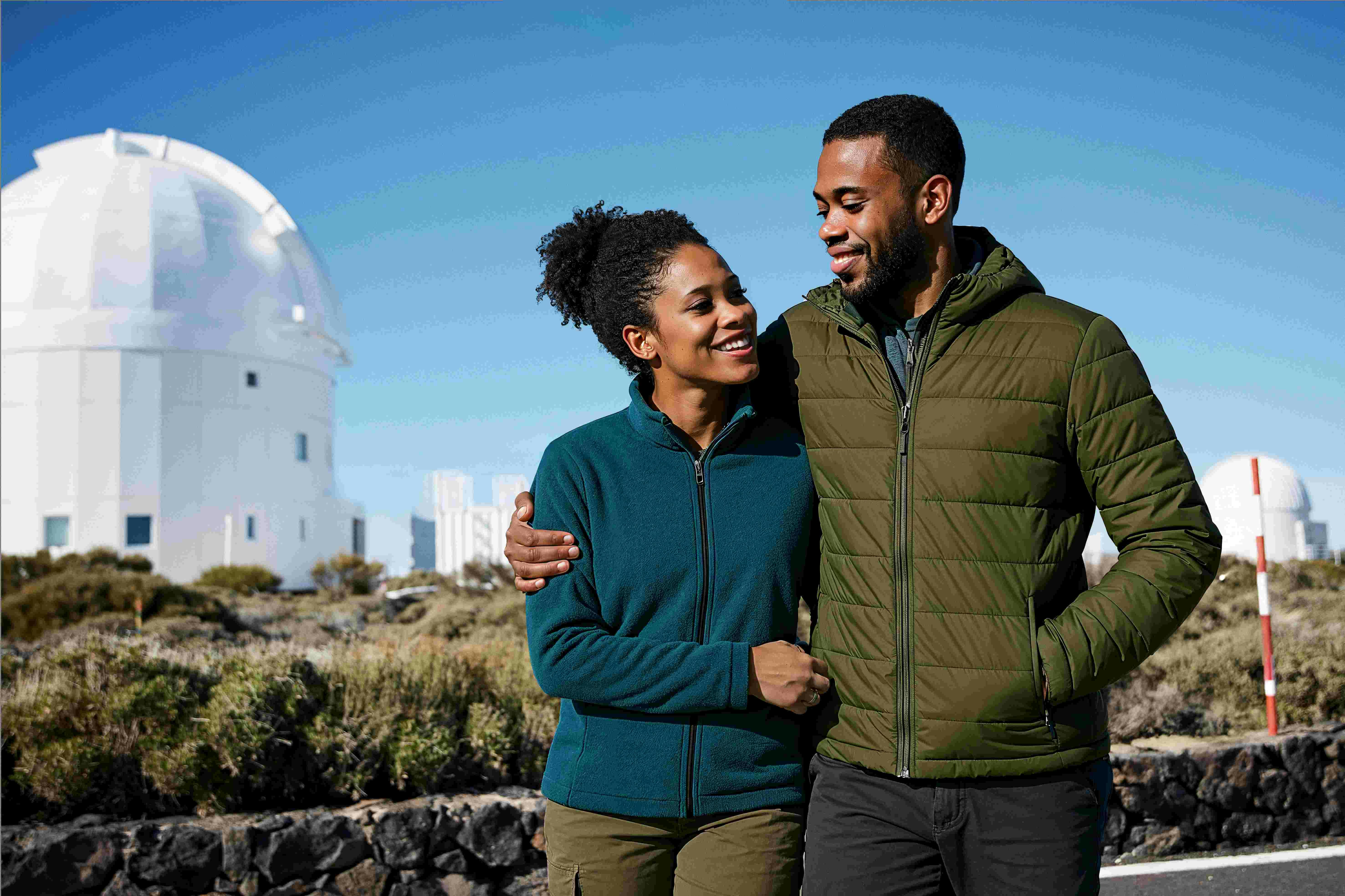 A smiling couple in jackets walks outdoors on a sunny day, with plants and buildings in the background.