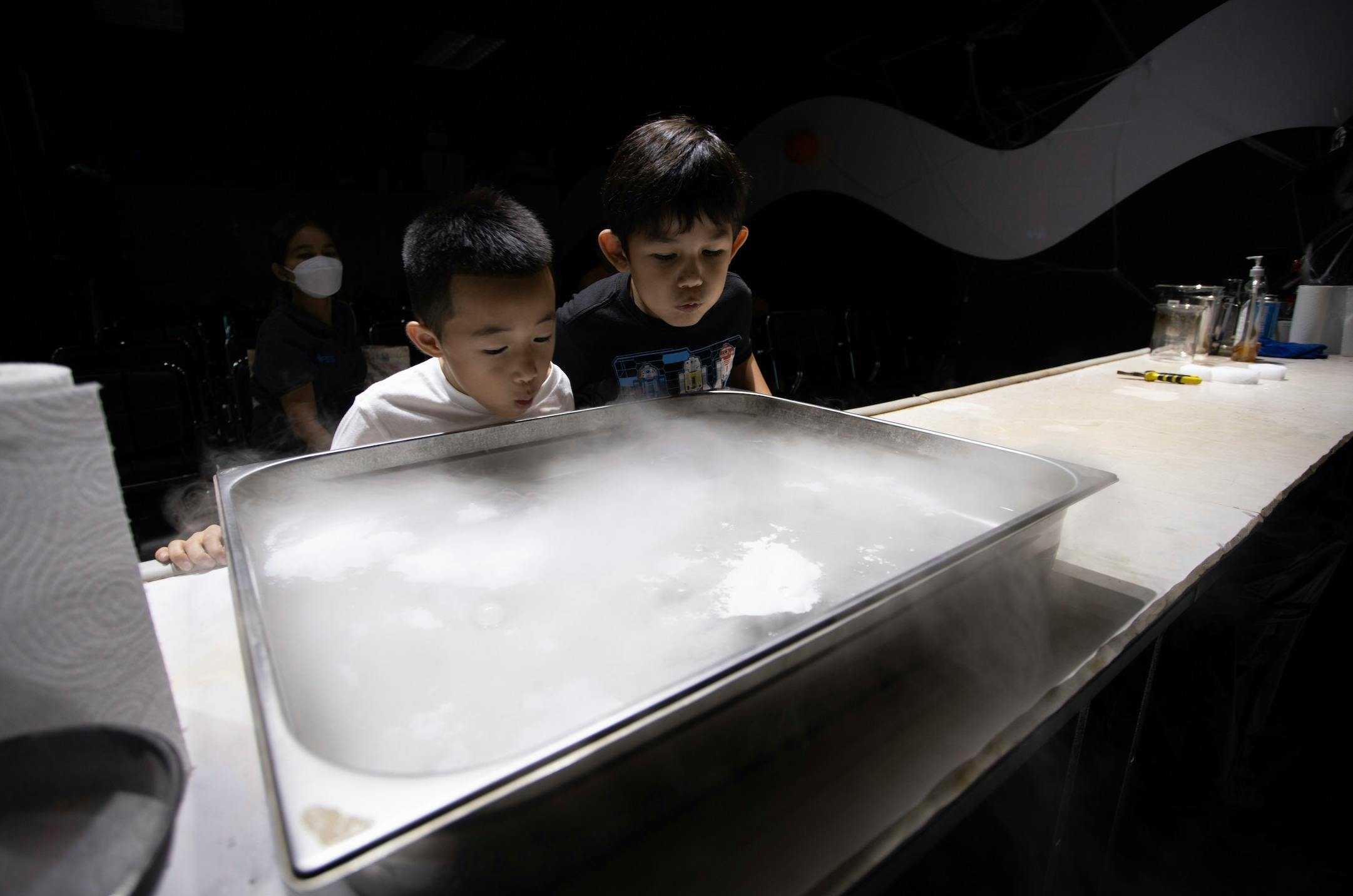 Two children leaning over a metal tray filled with mist and vapor, with a person wearing a mask in the background.