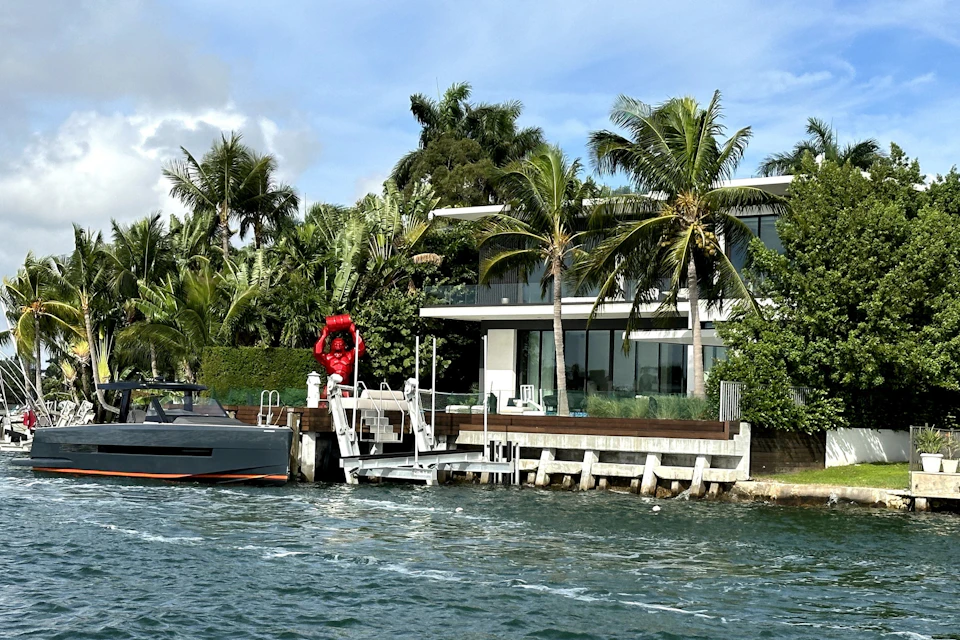 Modern waterside house with palm trees, boat dock, and red statue. Boat moored at dock. Sunny weather with a blue sky.