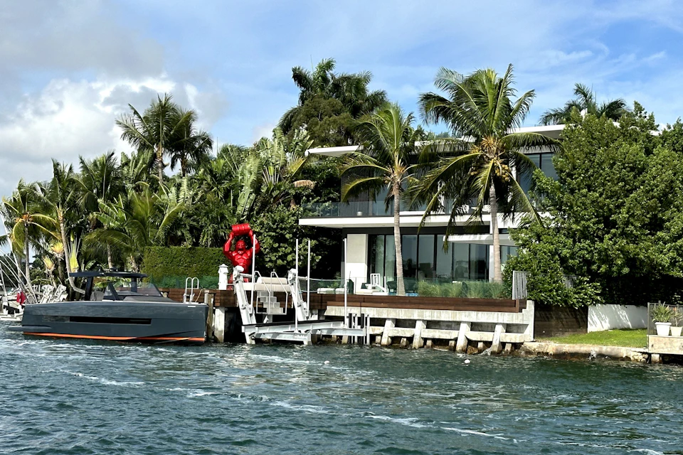 Modern waterside house with palm trees, boat dock, and red statue. Boat moored at dock. Sunny weather with a blue sky.