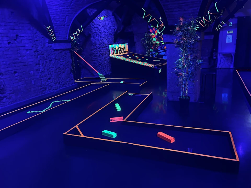 Indoor mini-golf course with neon obstacles, glowing decorations, and a "Pain Ball" sign, under blacklight.