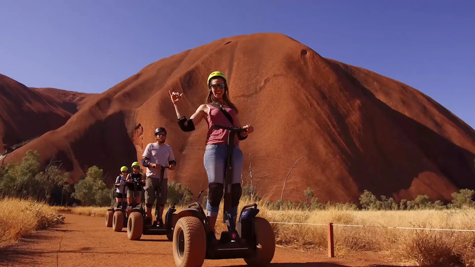 Four people riding Segways in a desert landscape with a large red rock formation in the background.