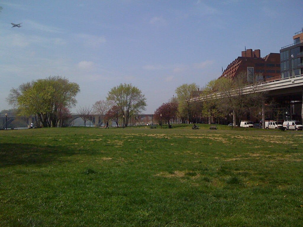 Grassy park with trees, a few people, a bridge, buildings, and an airplane in a partly cloudy sky.