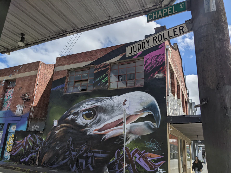 Street mural of an eagle's head on a brick building at the intersection of Chapel St. and Juddy Roller in an urban area.