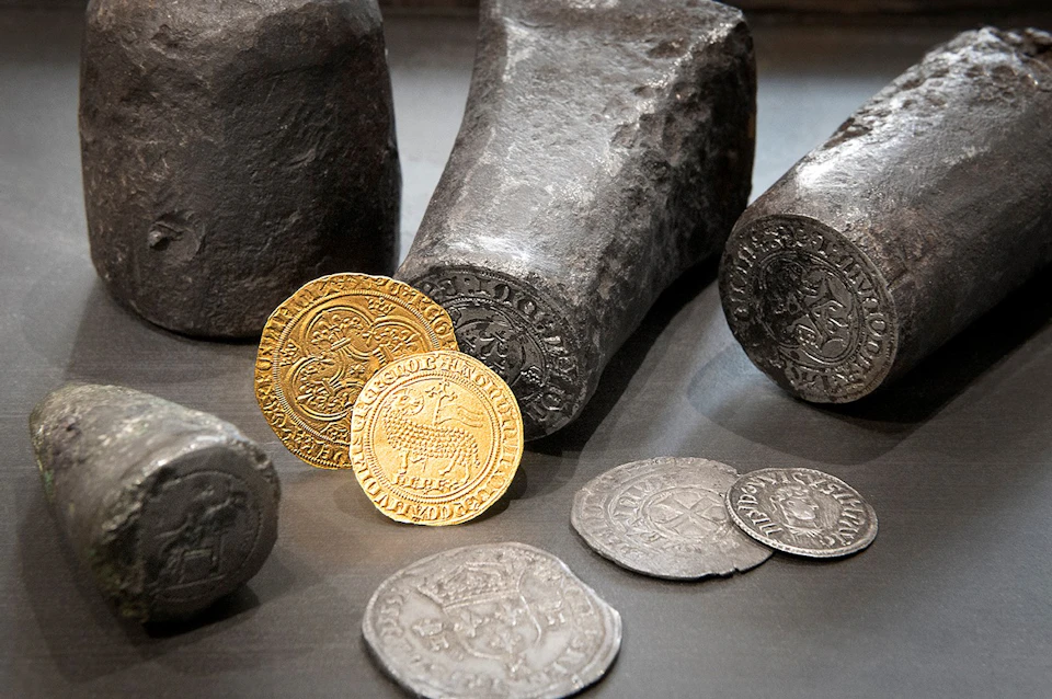 Old gold and silver coins placed in front of medieval coin stamps on a dark surface.