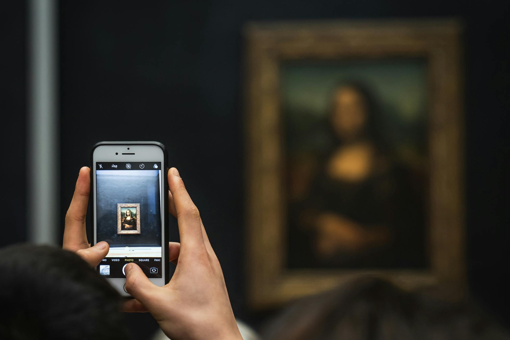 Personne tenant un smartphone, prenant une photo d'un tableau encadré de Mona Lisa. La foule et le tableau sont flous en arrière-plan.