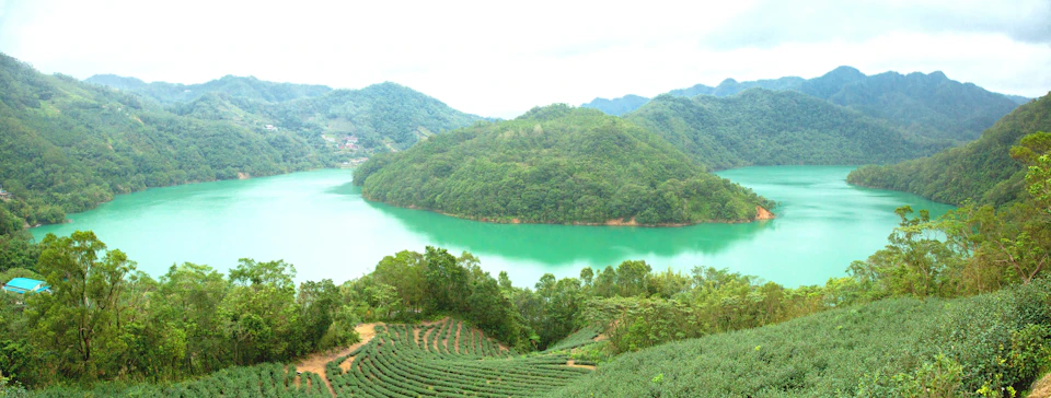 Lush green hills surround a jade-colored lake, with terraced fields in the foreground under a hazy sky.