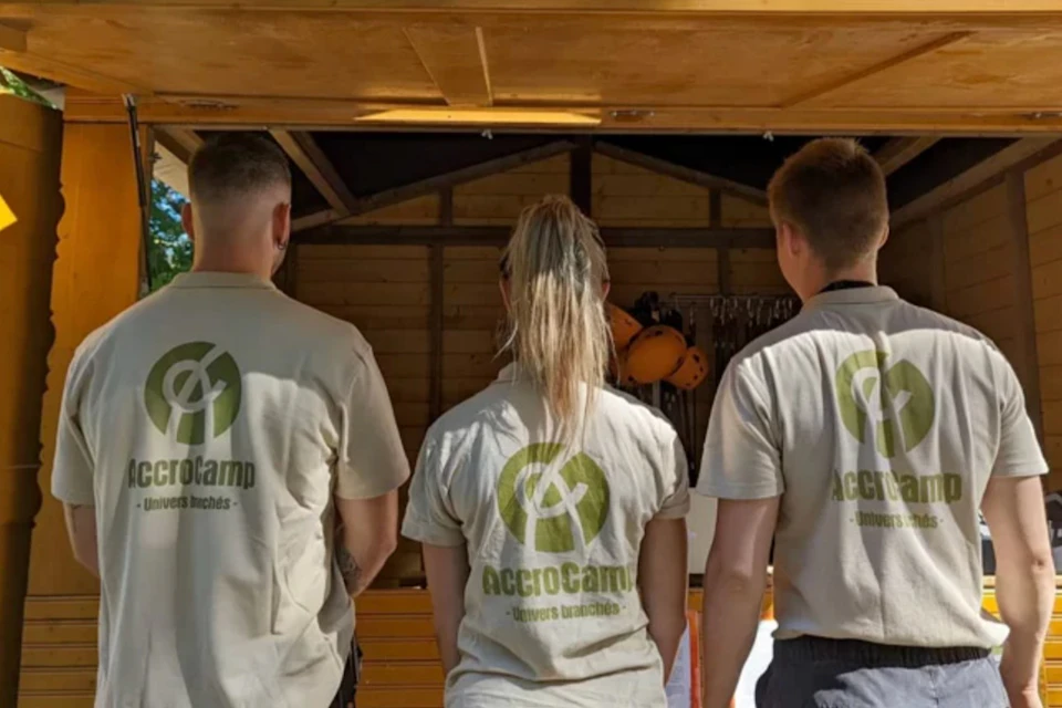 Three people with "AccroCamp" logos on their shirts stand facing away in front of a wooden structure with helmets inside.