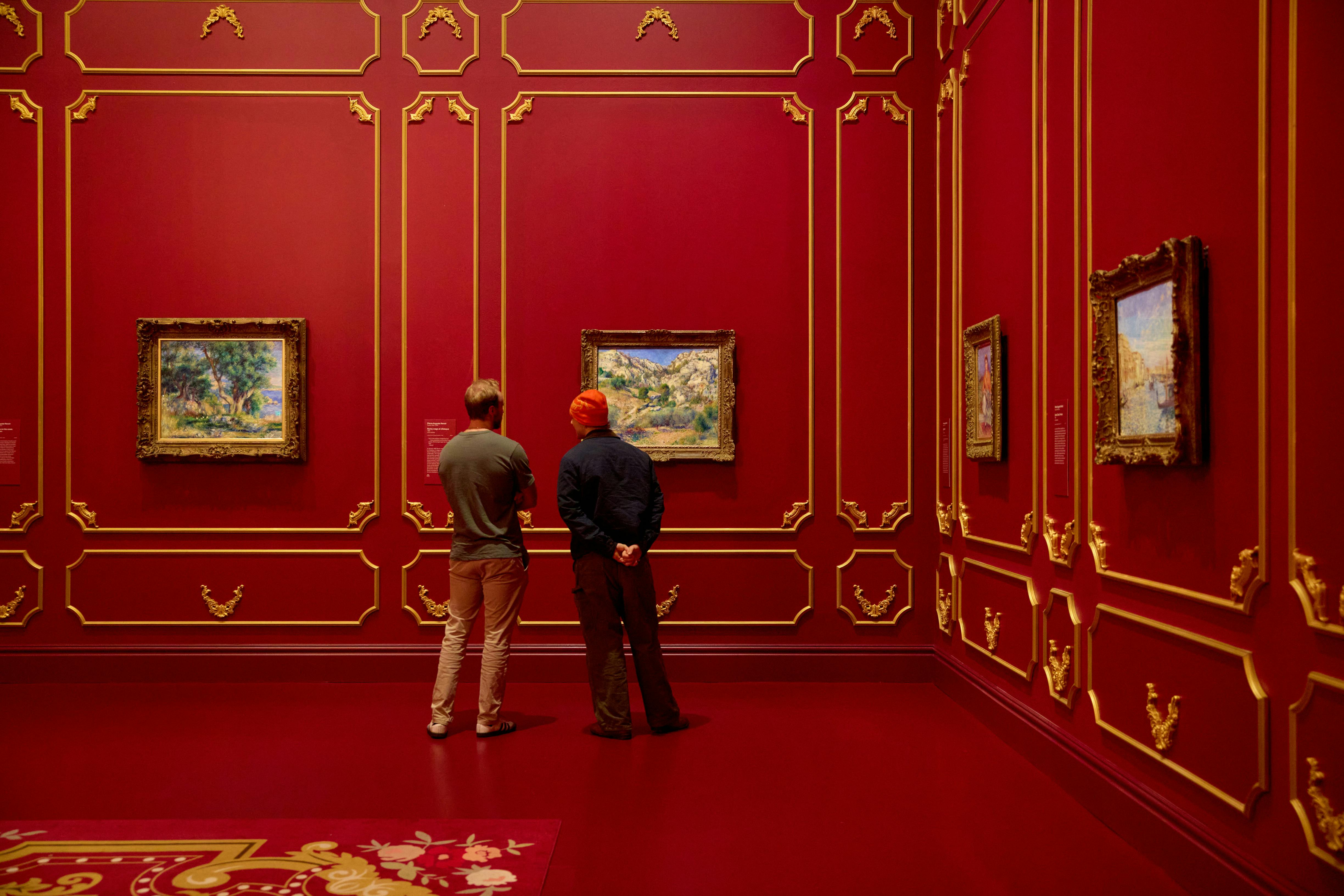 Dva lidé stojí těsně u sebe a prohlížejí si obrazy v galerii s červenými stěnami a zdobeným zlatým lemováním.